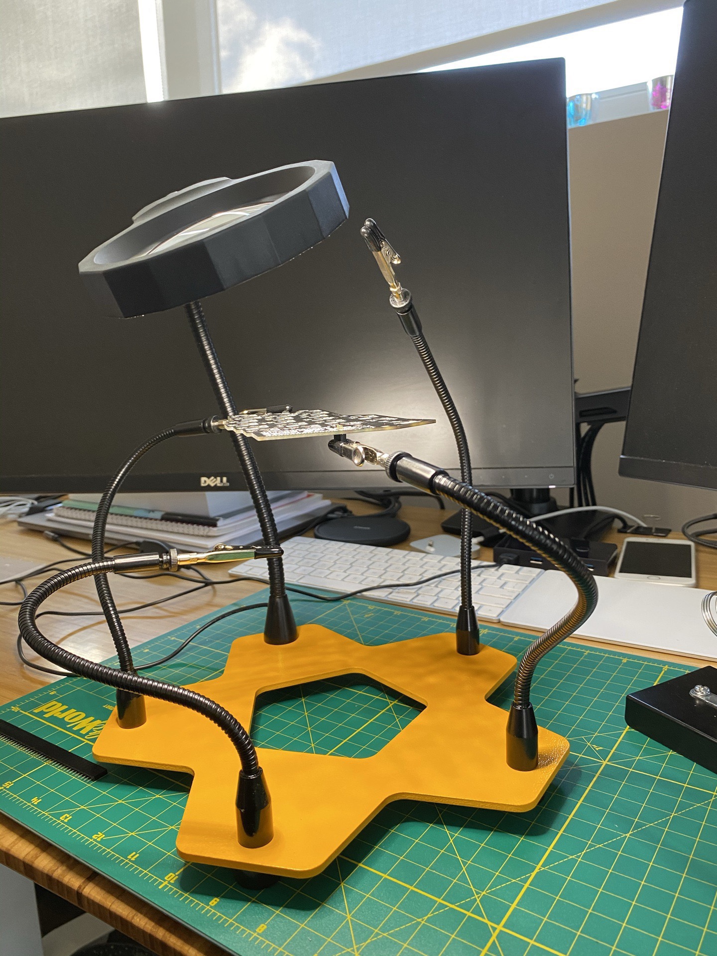 Workbench setup: helping-hands clamps mounted on a yellow base, a circular magnifying lamp, a green cutting mat, and Dell monitors with a window in the background.