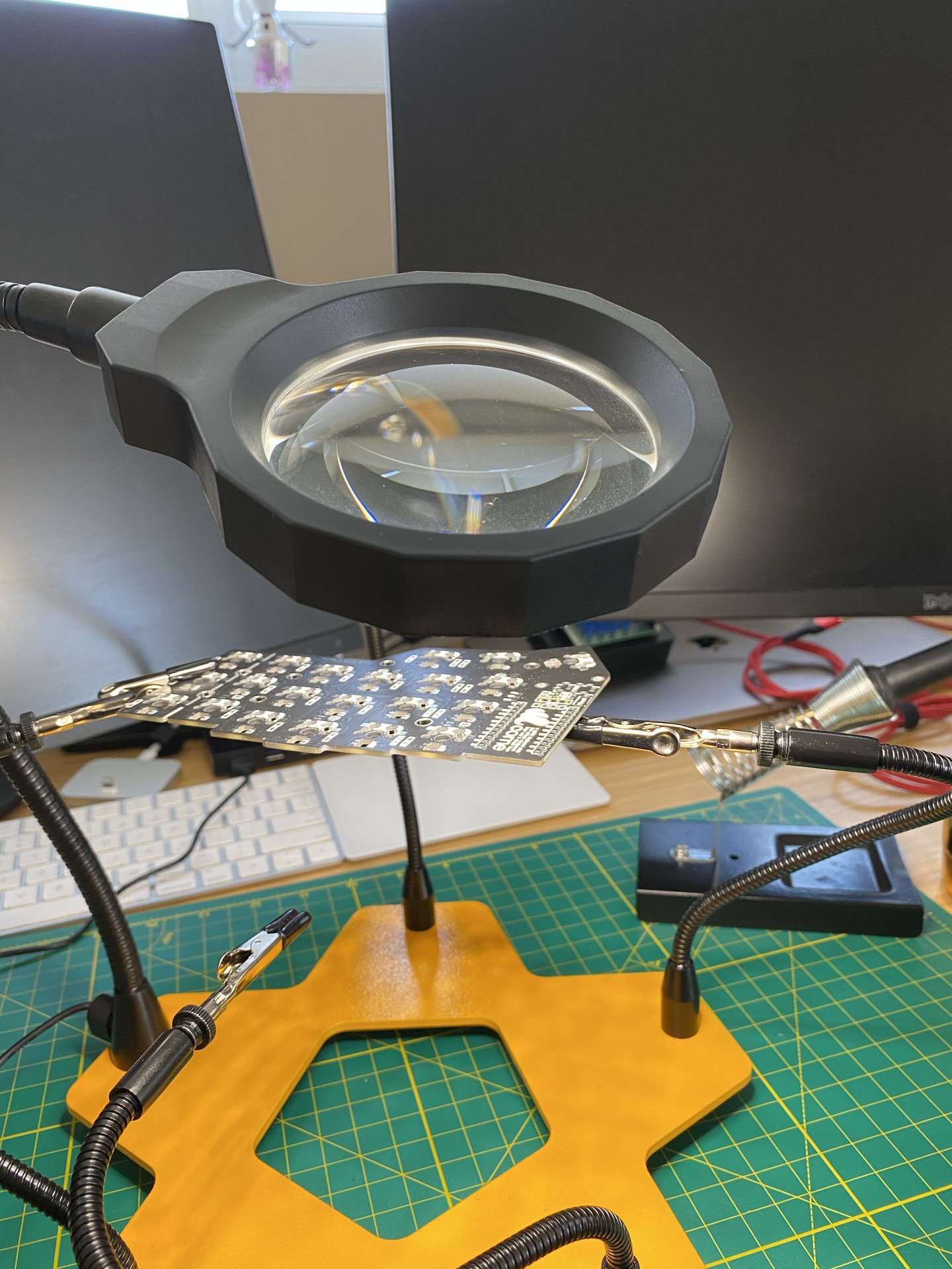 A bare Corne PCB suspended by the helping-hands clamps under the magnifying lamp, before any switches or keycaps were installed.
